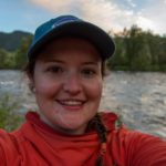 Image of Maggie, in a red hoodie and blue baseball hat in front of a river and trees.